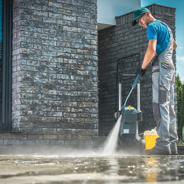 Pressure Washer Cleaning in Front of the House. Caucasian Men in His 30s Washing Concrete Bricks Driveway in Sunny Summer Day. Cleaning Around the House Concept.